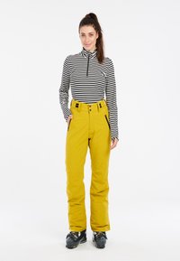Gelbe Schneehosen mit Reißverschlusstaschen und schwarzen Akzenten, kombiniert mit einem schwarz-weißen gestreiften Langarmshirt. Schwarze Skistiefel.