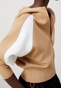 Beżowa bluza z kapturem o długości trzech ćwierć rękawów, z białym akcentem na ramieniu, ściągaczami na rękawach i zamkiem błyskawicznym.