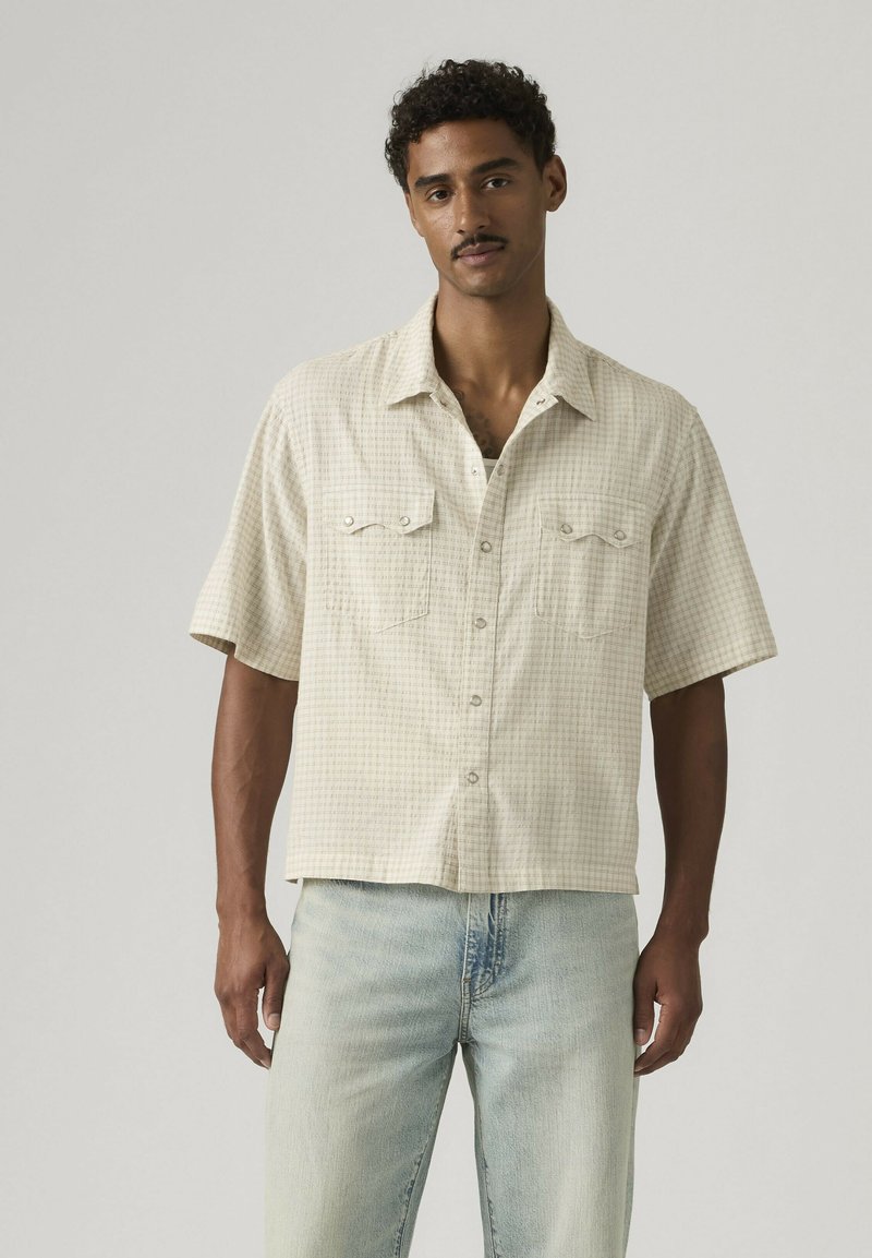 Hombre con camisa de cuadros beige de manga corta con bolsillos delanteros y vaqueros azul claro, de pie frente a un fondo gris liso.