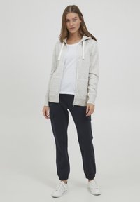 Hellgrauer Reißverschluss-Hoodie mit Kordelzügen, über einem weißen T-Shirt getragen, kombiniert mit marineblauen Cargo-Hosen und weißen Sneakern. Schlichtes Design.