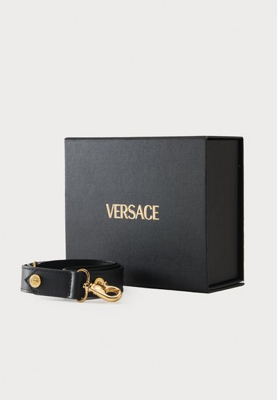 Čierny kožený remienok so zlatým kováním pred čiernou textúrovanou krabičkou, na viečku s logom „VERSACE“ zo zlata.