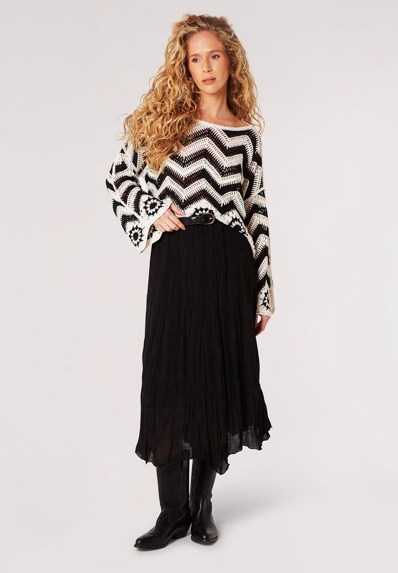 Häkelpullover mit schwarz-weißer Zickzackmuster, kombiniert mit einem langen, schwarzen plissierten Rock und schwarzen Lederstiefeln.