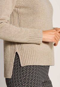 Beige Strickpullover mit gerippten Bündchen und Saum, getragen mit schwarz-weißen gemusterten Hosen, die seitliche Schlitzdetails zeigen.