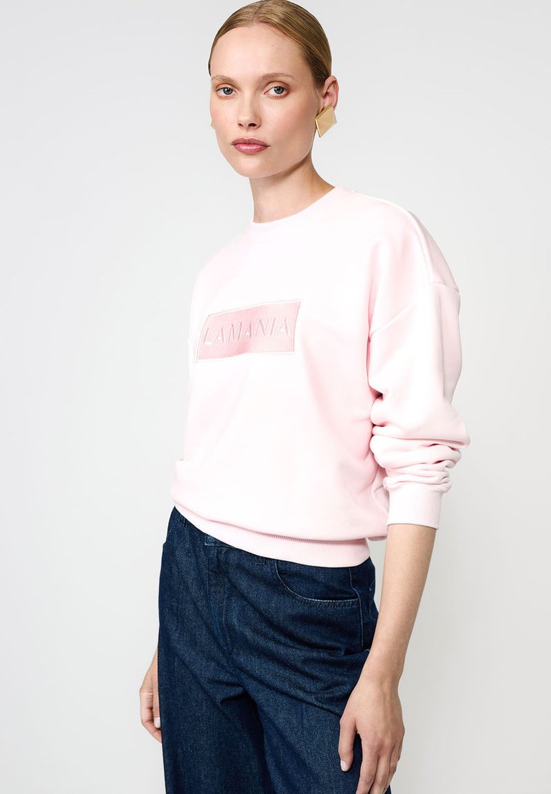 Heled roosa pusakas, millel on ümmargune kaelus ja reljeefne "LAMANIA" logo ristkülikukujulisel plaastril. Pehme kangas, mugav istuvus, soonikkoes varrukad.