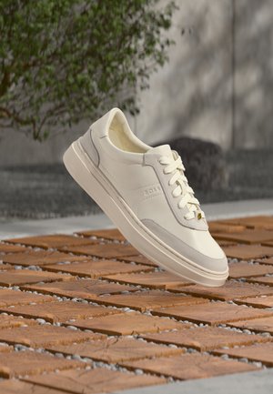 Białe sneakersy BOSS ze sznurowadłami unoszące się nad wzorzystą drewnianą i kamienistą powierzchnią, w tle zielony krzew i szara ściana.