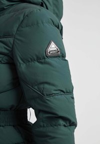 Chaqueta verde acolchada con textura acolchada, cintura elástica y un parche triangular plateado en el hombro.