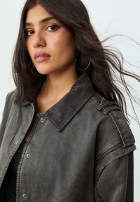 Grå denimjakke med corduroykrave, trykknaplukning og skulderepaulettes. Modellen har langt, bølget hår og bærer hoop øreringe.