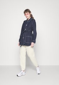 Dunkelblaue Denimjacke mit tailliertem Schnitt, vorderen Taschen, weißen Nähten, kombiniert mit cremefarbigen Fleecehosen und weißen Sneakers.
