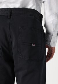 Navy cotton pants featuring rear pockets. Left pocket has a small logo tag with red, white, and blue accents. Smooth texture, standard fit.