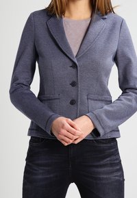 Marinblå blazer i en texturerad tyg, med slagknappar, tre knappar och två framfickor, kombinerad med en grå överdel och mörka jeans.