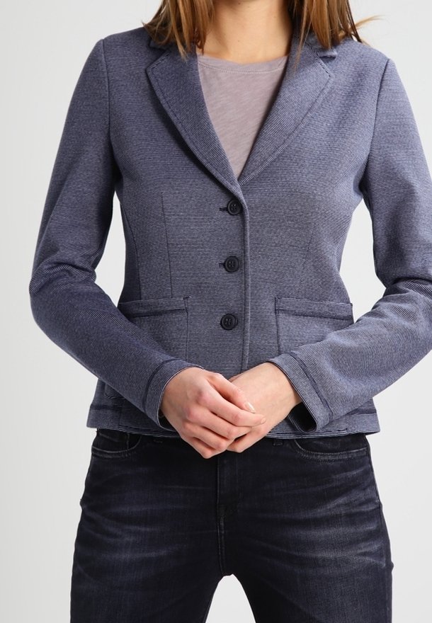 Marinblå blazer i en texturerad tyg, med slagknappar, tre knappar och två framfickor, kombinerad med en grå överdel och mörka jeans.