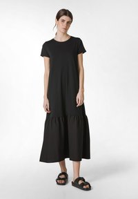 Černé bavlněné maxi šaty se přiléhavým vrškem a vrstveným volánovým lemem. Spárováno s černými sandály s detailním sponou. Jednoduchý design.