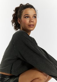 Schwarzer Pullover aus strukturiertem Material, cropped Schnitt und weiten Ärmeln. Das Model trägt einen schwarzen Lederrock. Neutraler Hintergrund.