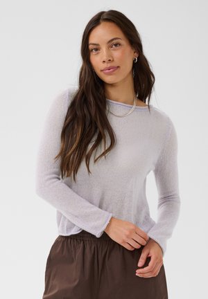 Femme aux longs cheveux bruns portant un pull violet clair à manches longues, un pantalon marron, des boucles d’oreilles créoles et un collier chaîne, debout devant un fond uni.