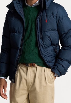 Hombre con chaqueta acolchada azul marino sobre suéter verde de punto con trenzas, pantalones beige, cinturón marrón y reloj de pulsera, con la mano en el bolsillo.