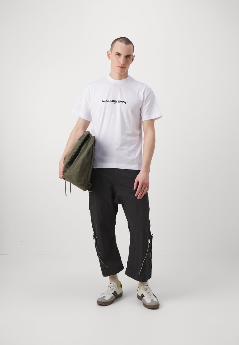 T-shirt bianco con testo nero, pantaloni cargo neri con tasche con cerniera, borsa a tracolla verde e scarpe da ginnastica multicolori; outfit casual.