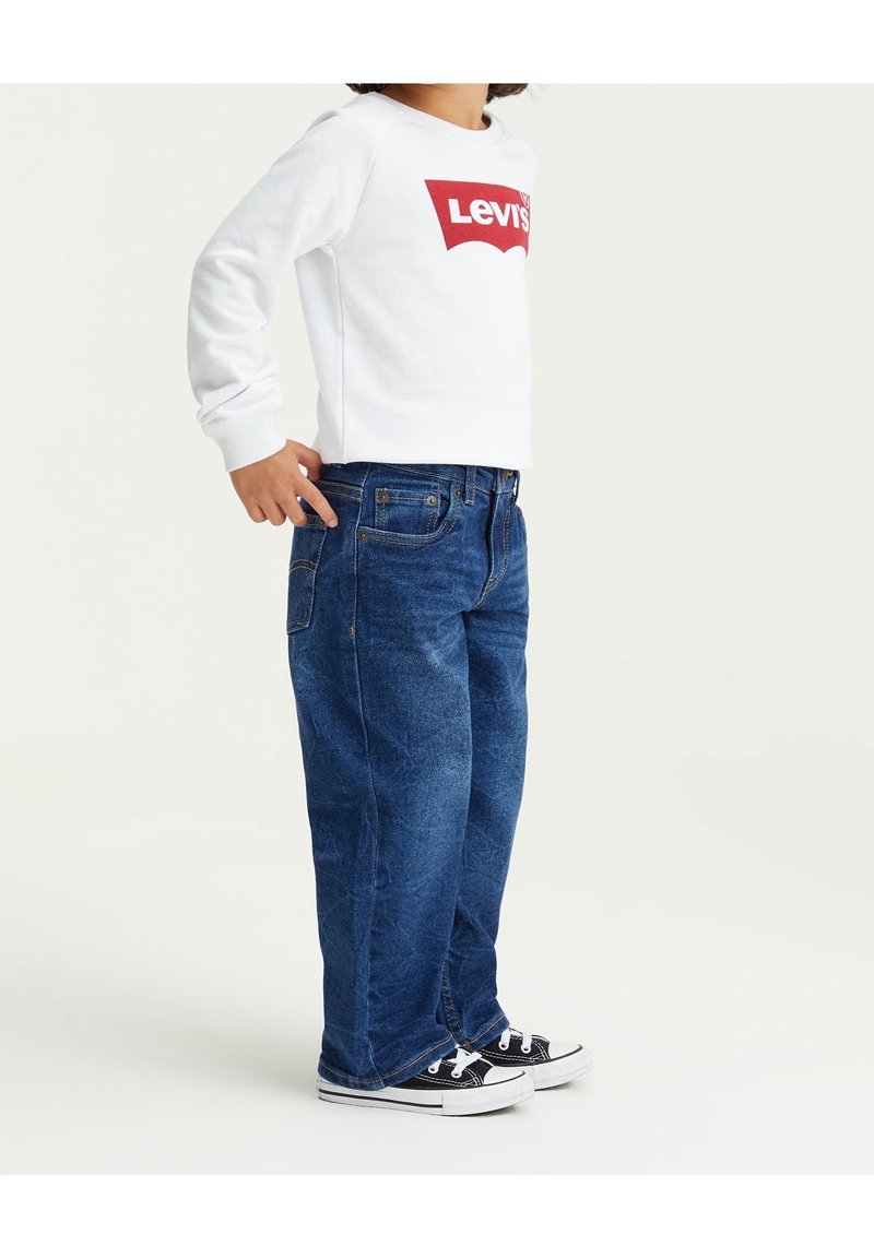 Sudadera blanca con logo rojo de Levi's, combinada con jeans de denim azul y zapatillas negras, mostrando un ajuste relajado y un diseño clásico.