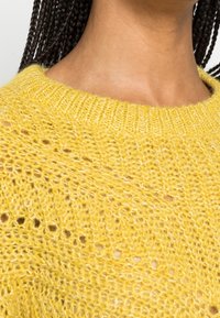 Nahaufnahme einer Person mit geflochtenen Haaren, die einen strukturierten gelben Strickpullover mit rundem Kragen trägt.