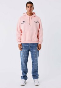 Lichtroze hoodie met een voorzak, trekkoorden en een marineblauw logo. Gecombineerd met blauwe spijkerbroeken met een herhalend logo-patroon. Witte sneakers.
