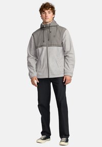 Grey and light grey hooded jacket with a front zipper, elastic cuffs, and a patterned stripe across the chest. Paired with black pants and sneakers.