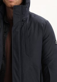 Giacca invernale blu scuro, con cappuccio, texture liscia, chiusura con zip e dettagli di design angolari. Indossata sopra un maglione lavorato a maglia marrone.