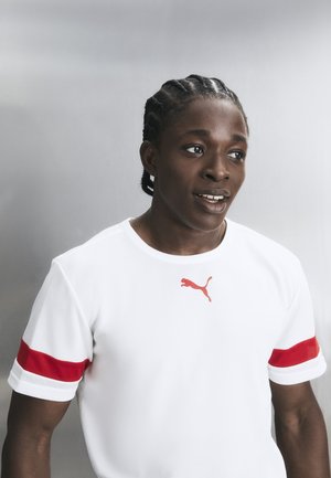 Νεαρός άνδρας με πλεξούδες στα μαλλιά, φοράει λευκό μπλουζάκι Puma με κόκκινες ρίγες στα μανίκια και κοιτάζει λίγο προς τα πλάγια με γκρι φόντο.