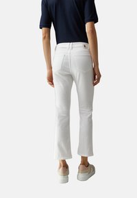 Jean blanc évasé avec une texture lisse, deux poches arrière, et un détail logo à la ceinture, associé à des baskets beige clair.