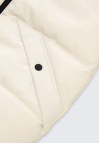 Close-up van crèmekleurige gewatteerde stof met diagonale stiksels en een zwarte drukknoop op een zakflap.