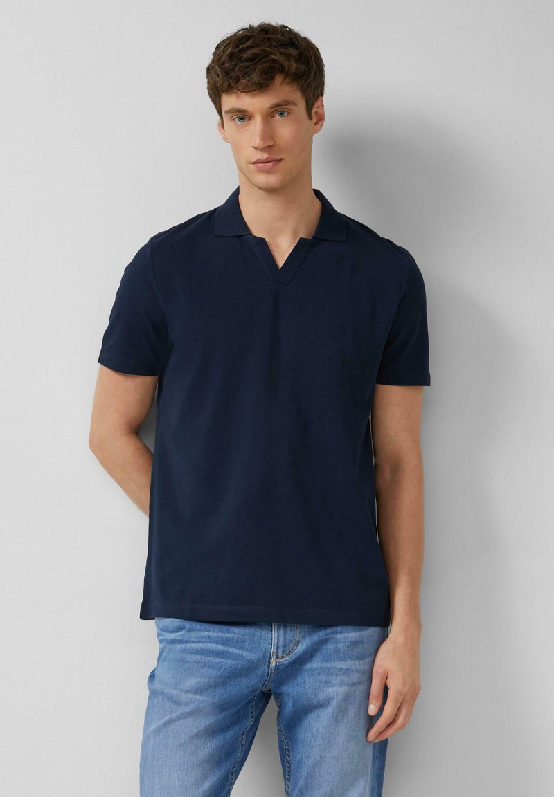 Ein junger Mann mit kurzen lockigen Haaren trägt ein marineblaues Polohemd mit kurzen Ärmeln und hellblaue Jeans, steht vor einer weißen Wand.