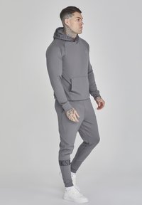 Szürke kapucnis felső és hozzá illő joggingnadrág sima anyagból, elülső zsebbel, bordázott mandzsettákkal és logóval a nadrágon. Fehér sportcipők.
