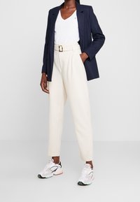 Granatowa marynarka, biały t-shirt w serek, beżowe spodnie z wysokim stanem z paskiem oraz białe sneakersy z wielokolorowymi akcentami. Gładkie tekstury.