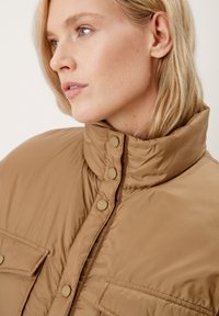 Blonde Frau, die nach links schaut und eine hochkragen, beigefarbene, gepolsterte Jacke mit Knopfverschlüssen und Klappentaschen trägt.