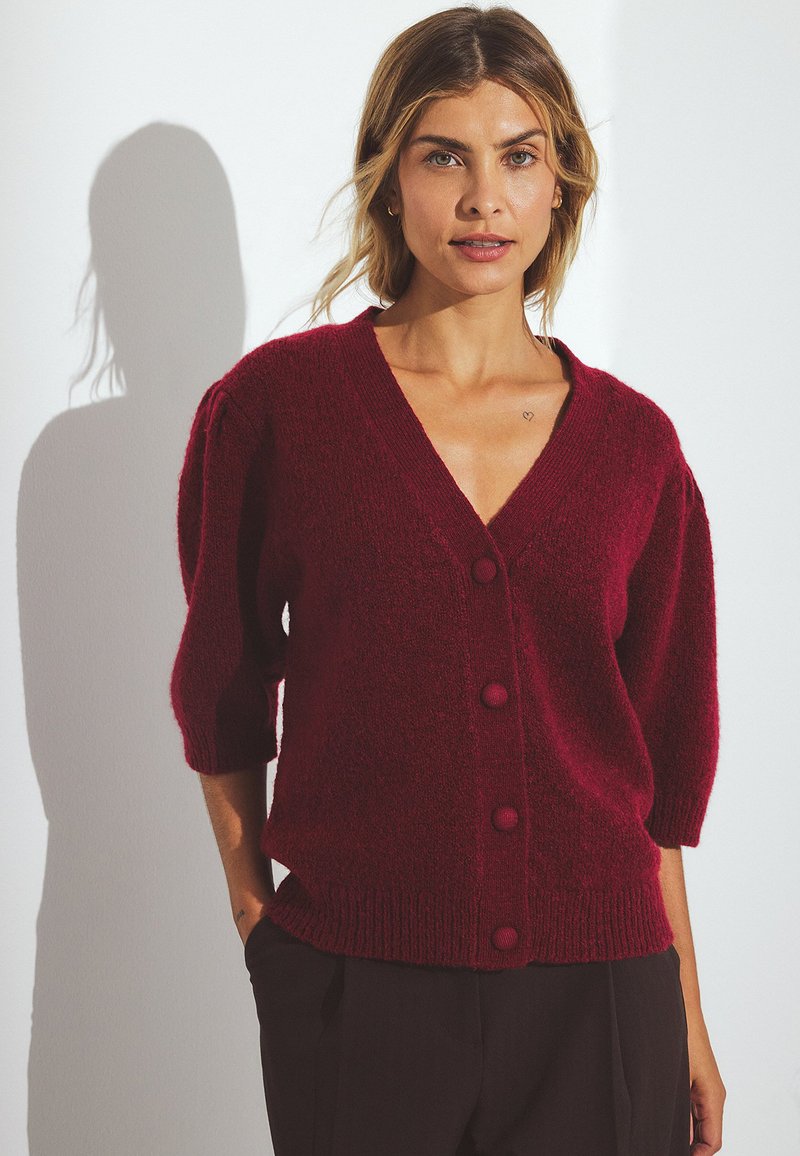 Burgundijas adīts kardigāns ar V veida kakla izgriezumu, īsām pūkainām piedurknēm un piecu pogu aizdari. Mīksta tekstūra, ribota apakšmala un brīvs piegriezums.