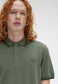 Polo verde con tejido texturizado, tapeta de doble botón y acentos de collar a rayas. Presenta un logo en el lado izquierdo del pecho.
