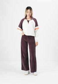 Vrouw staand in een wit en paars ritsjack met oranje strepen, bruine wijde corduroy broek en witte sneakers tegen een eenvoudige witte achtergrond.