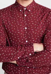 Camisa de botones color burdeos con patrón de plumas blancas, de mangas largas y puños con botones. Fabricada con un tejido ligero, presenta un cuello clásico.