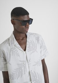 Camisa de manga corta, de color gris claro, con un sutil patrón de helechos y botones blancos. Unas gafas de sol cuadradas oscuras completan el conjunto.