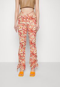 Calças coloridas com padrões em espiral de vermelho, laranja e verde; feitas de um tecido macio; detalhe de cordão ajustável nos tornozelos; combinadas com saltos laranjas.