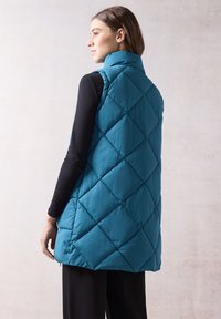 En teal-färgad quiltad puffer-väst med hög krage och diamantformad sömnad, bärs över en svart långärmad topp. Slät textur.