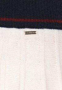 Neulotut neulepaidat, joissa on tummansininen yläosa ja kerma vaalean alaosa, joissa on pystysuora ribbitekstuuri. Sisältää metallisen logovivahteen.