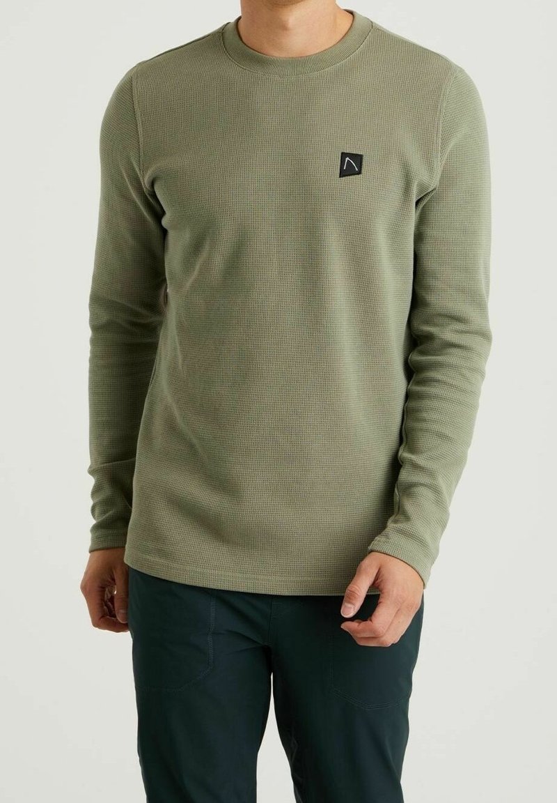 Homme portant une chemise texturée vert olive à manches longues avec col rond et un pantalon foncé, debout devant un fond clair uni.