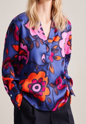 Kobieta ubrana w luźną bluzkę z długim rękawem, ozdobioną jaskrawymi różowymi, pomarańczowymi i czerwonymi kwiatowymi wzorami na niebieskim tle.