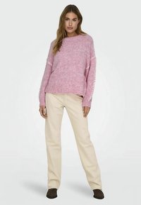 Różowy dziany sweter o luźnym kroju i szerokich rękawach, w zestawieniu z beżowymi spodniami i brązowymi butami. Materiał o fakturze z delikatnymi zdobieniami.