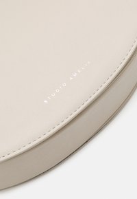 Borsa rotonda in pelle beige con superficie testurizzata. Presenta cuciture sottili e la scritta in rilievo "STUDIO AMELIA" in bianco. Nessun hardware visibile.