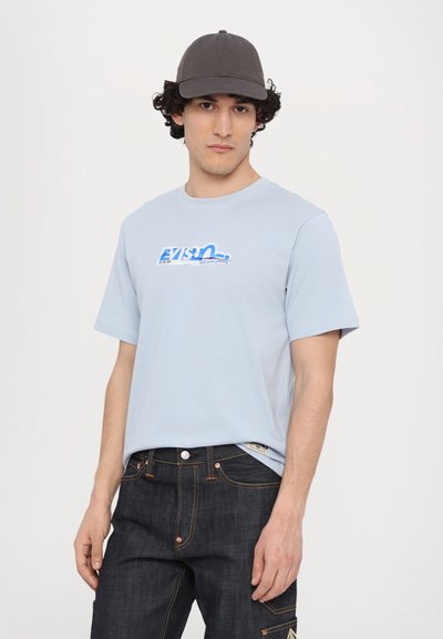 Light blue short-sleeve t-shirt with a blue graphic logo, paired with dark denim jeans featuring orange stitching and a side pocket detail.