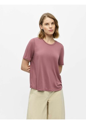 Kvinde med kort blondt hår iført en mauve kortærmet top og beige vidde bukser, stående med hænderne bag ryggen.