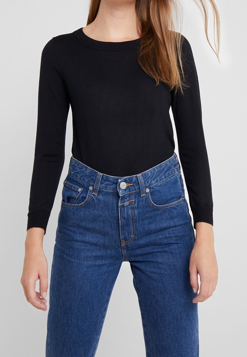 Femme portant un haut noir à manches longues rentré dans un jean bleu taille haute avec poches avant et fermeture à boutons.