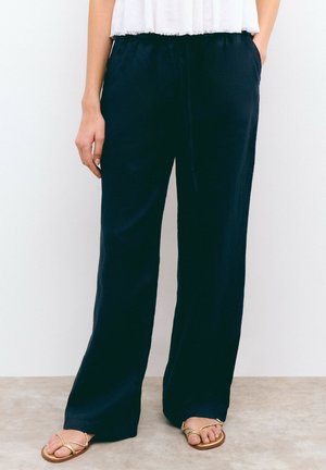 Pantalons marine à jambes larges en tissu doux, dotés d'une taille élastique avec cordon de serrage. Portés avec des sandales à brides dorées.