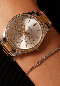 Gold- und Silberuhr mit rundem Ziffernblatt, drei Nebenzifferblättern, einer mit Kristallen besetzten Lünette und einem metallischen Armband. Daneben liegt ein schlanker Silberarmreif.