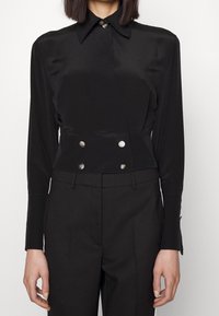 Osoba oblečená v černé košili s dlouhým rukávem a širokým límcem, vybavené čtyřmi stříbrnými knoflíky v pase, doplněná černými přizpůsobenými kalhotami.